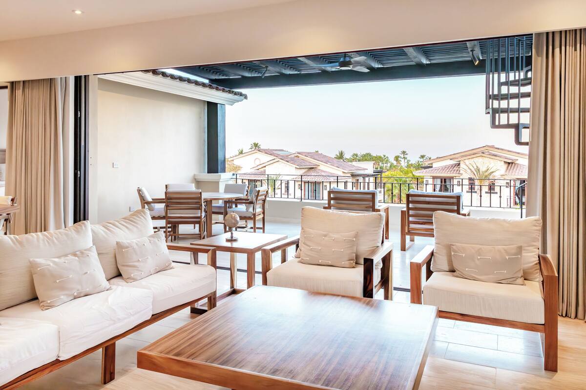 A modern lounge area with a sofa, chairs, and a coffee table. There is a view of a building with a tiled roof and palm trees in the background.