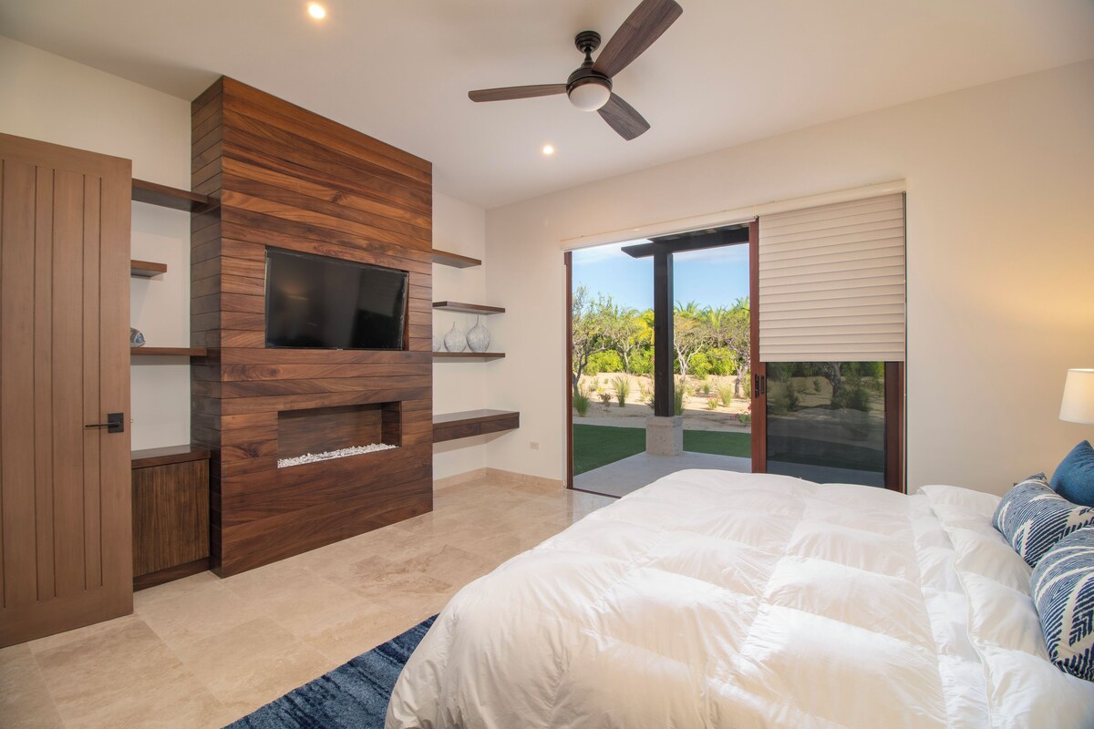 A bedroom with a large bed, a TV mounted on a wooden wall, a ceiling fan, and a sliding glass door leading to a balcony.