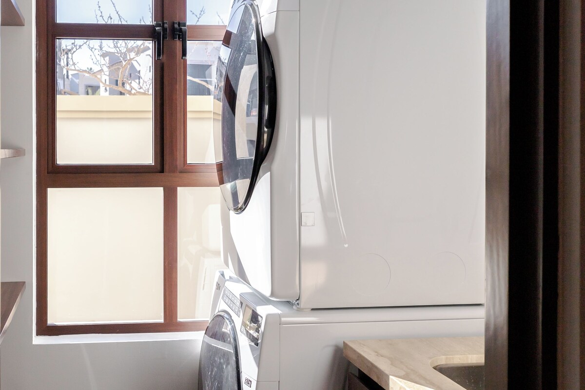 Une petite buanderie équipée d’un lave-linge et d’un sèche-linge, d’une fenêtre avec des stores et d’un plan de travail en bois.