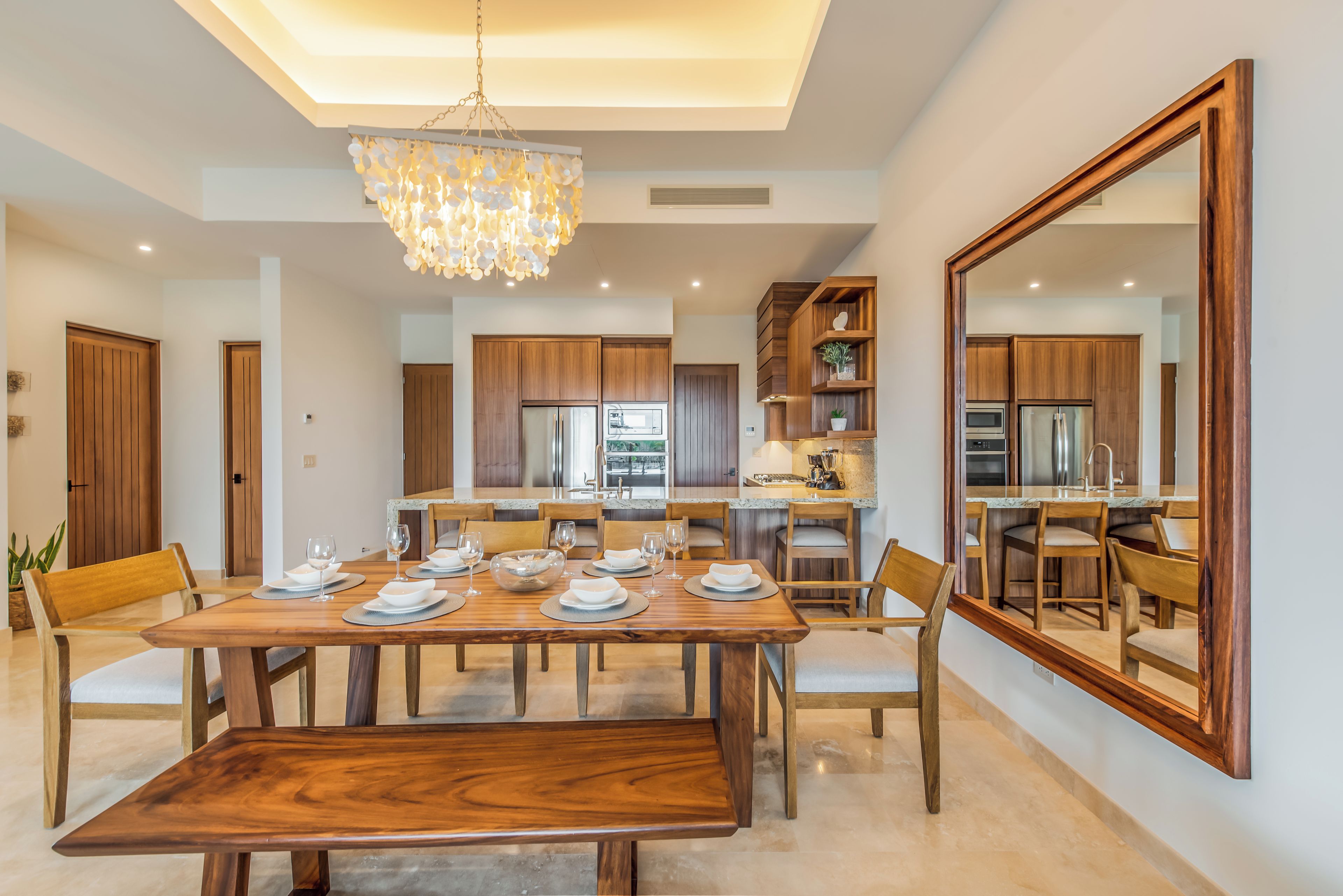 A modern dining area with a wooden table set for six, surrounded by chairs. A large mirror reflects the room, and a chandelier hangs from the ceiling.