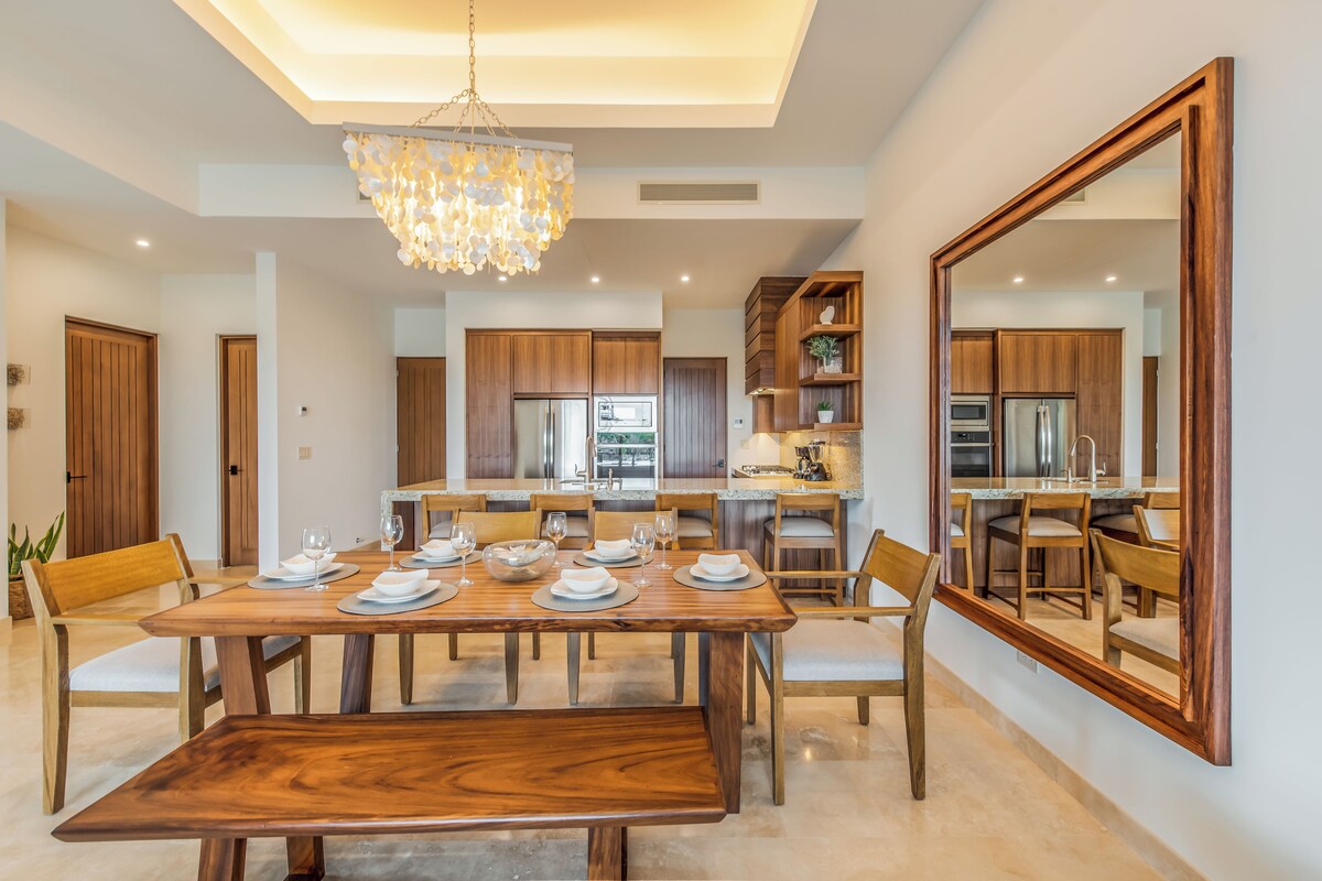 A modern dining area with a wooden table set for six, surrounded by chairs. A large mirror reflects the room, and a chandelier hangs from the ceiling.