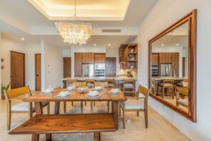 A modern dining area with a wooden table set for six, surrounded by chairs. A large mirror reflects the room, and a chandelier hangs from the ceiling. - Bohari (San José del Cabo)