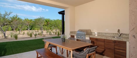 Cocina exterior con parrilla, mesa de comedor y sillas, rodeada de un jardín ajardinado.