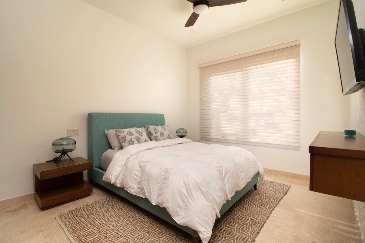 A bedroom with a bed, nightstands, a ceiling fan, a window with blinds, and a TV on a stand.