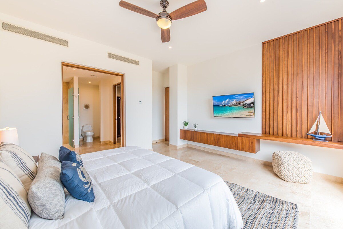A modern bedroom with a large bed, a ceiling fan, a wooden panel wall, a mounted TV, and a small sailboat decoration.
