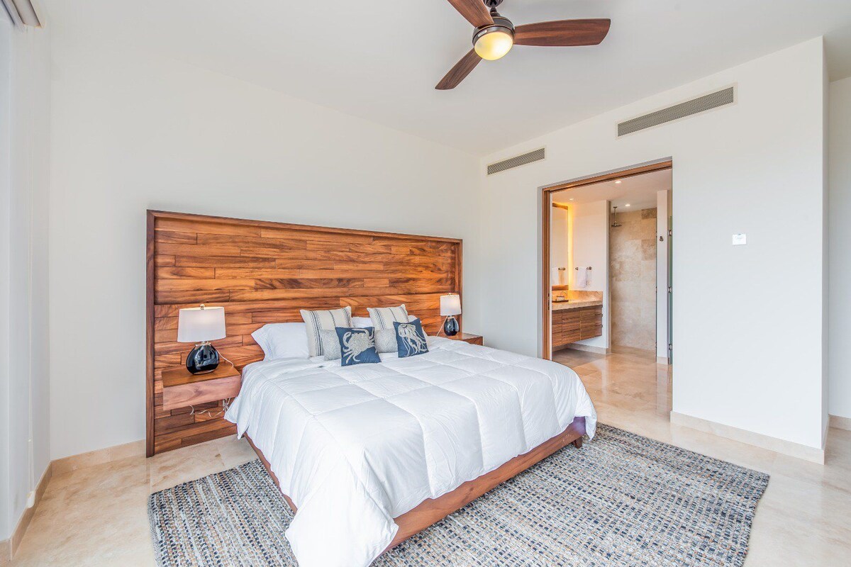 A bedroom with a wooden headboard, a large bed with white bedding, two bedside tables with lamps, and a ceiling fan.