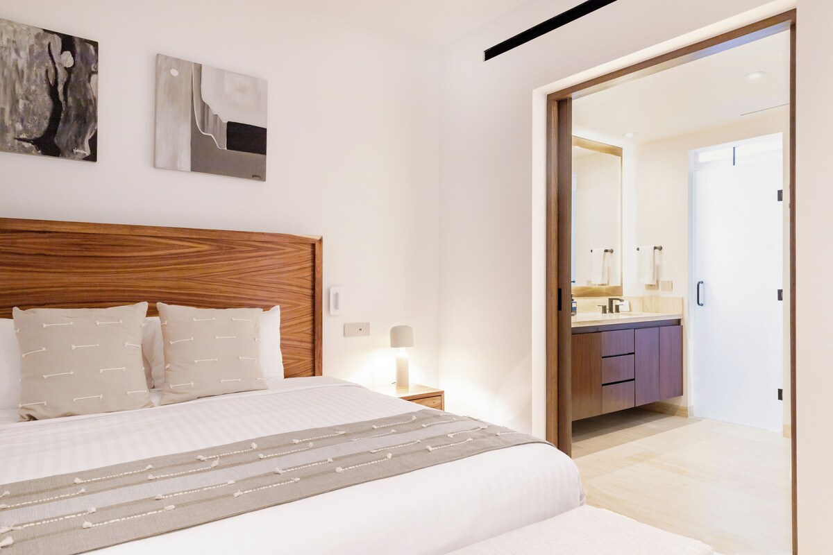A modern hotel room with a wooden headboard, a bed with white linens, a bedside table with a lamp, and a bathroom visible through an open door.