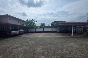 A parking area with a concrete surface, a covered carport, and a building with a flat roof. - Phaiboon Hotel & Resort By Zuzu (Pak Chong)