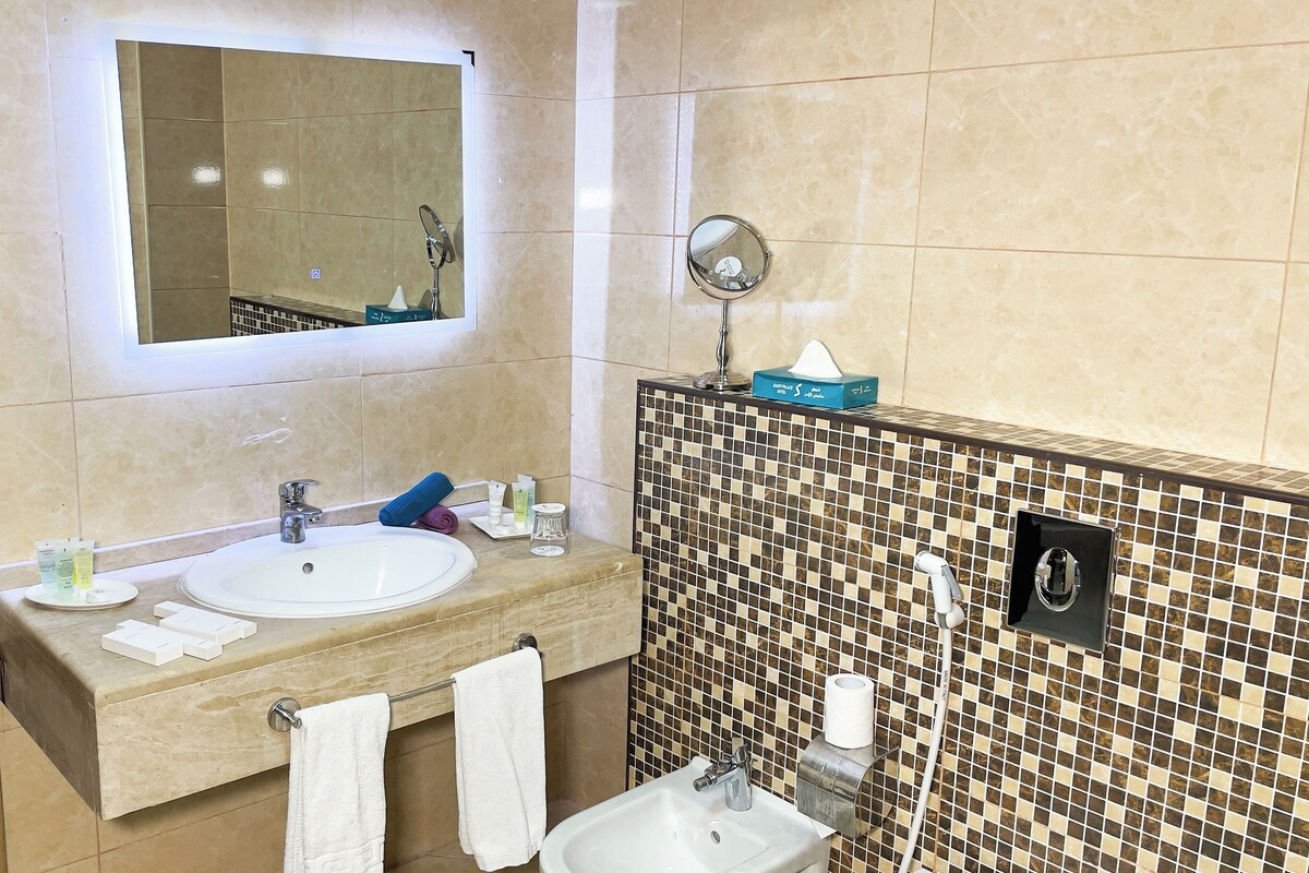 A bathroom with a large mirror, two sinks, a small sink, a towel rack, and a tiled shower wall.