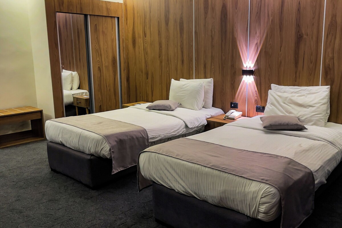 A hotel room with two beds, a wooden wardrobe, a bedside table with a lamp, and a small wooden bench.