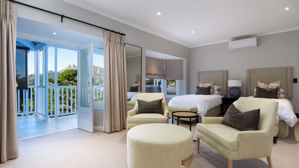 A spacious bedroom with a large bed, two armchairs, a round ottoman, and a small table. A balcony with a view of greenery and a mountain is visible through an open door. - Bay Haven Collection, Hout Bay (Cape Town)