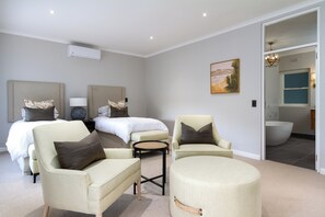 A modern hotel room with two beds, a sitting area with chairs, a round ottoman, and a view of the bathroom. - Bay Haven Collection, Hout Bay (Cape Town)