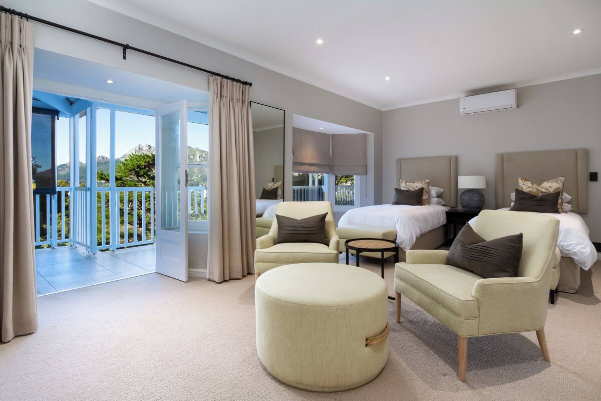 A spacious bedroom with a large bed, two armchairs, a round ottoman, and a small table. A balcony with a view of greenery and a mountain is visible through an open door.