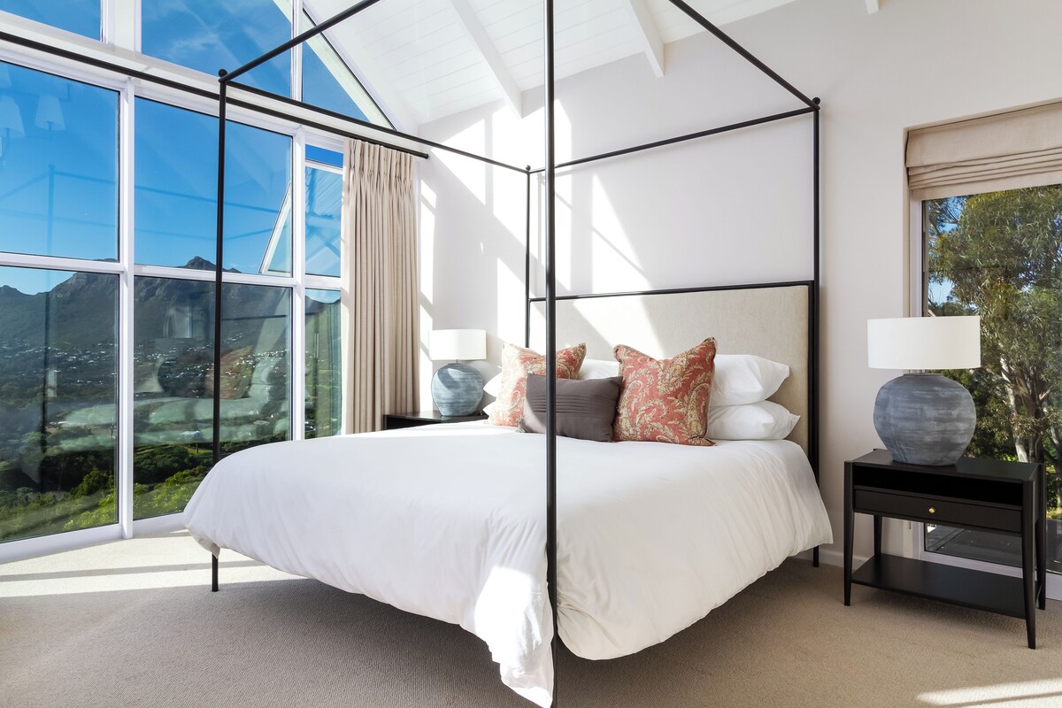 A modern bedroom with a large bed, a glass wall, and a view of the outdoors.