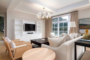 Un salon moderne comprenant un canapé, des fauteuils, une table basse, un téléviseur posé sur un meuble blanc et une grande fenêtre avec des rideaux.