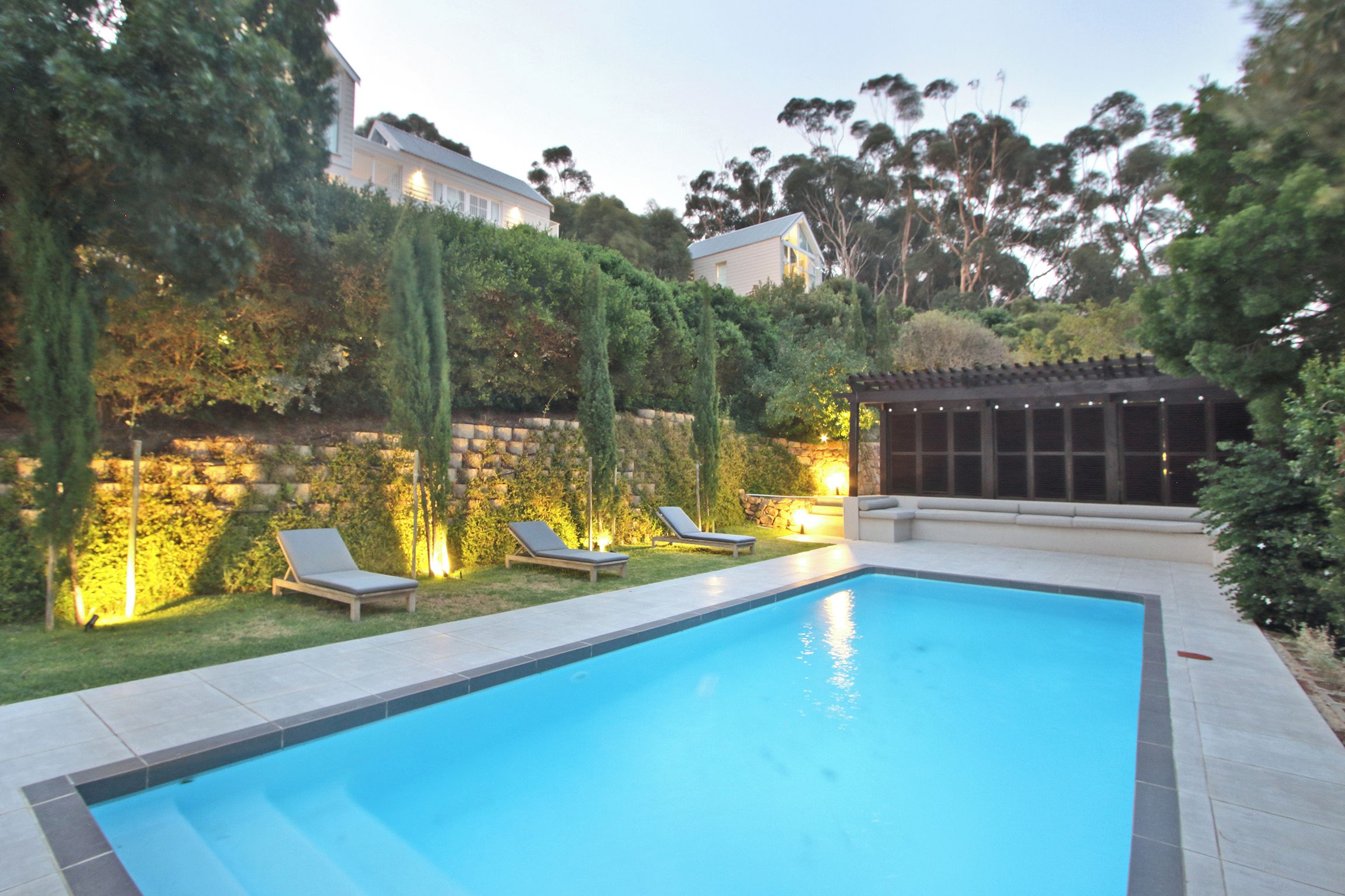 Ein rechteckiger Swimmingpool mit zwei Liegestühlen und einer Pergola mit Beleuchtung, umgeben von einer Steinmauer und Grünpflanzen.