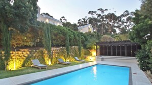 A rectangular swimming pool with two lounge chairs and a pergola with lights, surrounded by a stone wall and greenery. - Bay Haven Collection, Hout Bay (Cape Town)