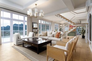 A spacious living room with a large window, a dark coffee table, white sofas, and a staircase leading to an upper level.