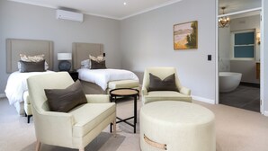 A modern hotel room with two beds, a sitting area with chairs, a round ottoman, and a view of the bathroom. - Bay Haven Collection, Hout Bay (Cape Town)