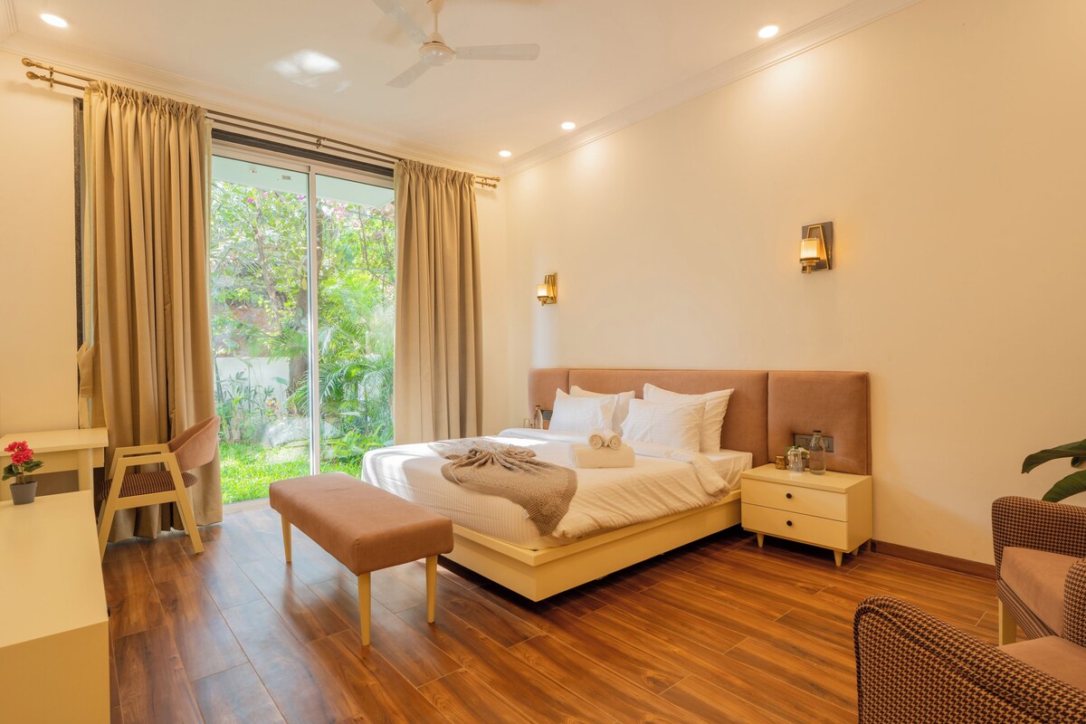 A modern bedroom with a large bed, bedside tables, a bench, and a view of greenery through the window.