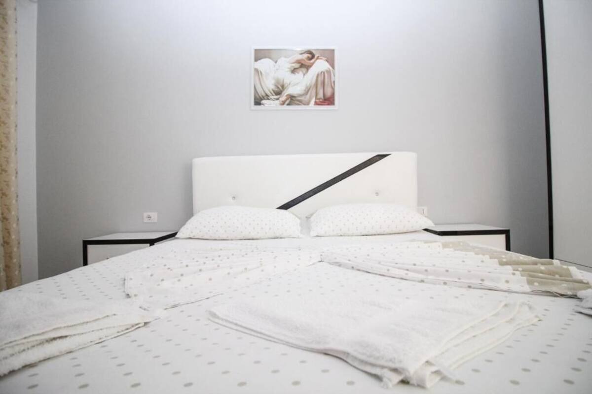 A neatly made bed with white bedding and pillows, a framed artwork above the headboard, and a black bedside table.