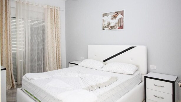 A bedroom with a bed, a nightstand, a drawer, a window with curtains, and a wall-mounted picture. - Hotel Artur 2 (Sarandë)
