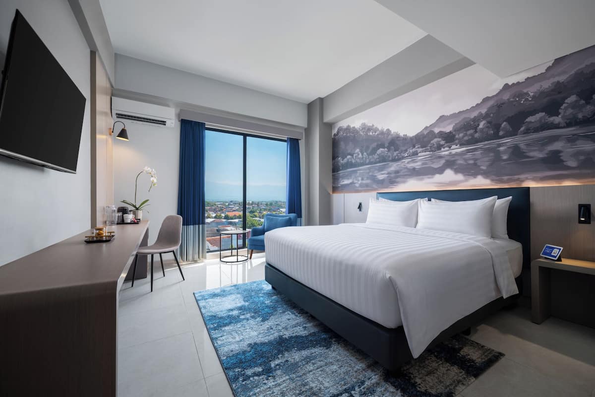 Habitación de hotel moderna con una cama grande, un escritorio, una silla, una mesita y un ventanal con vistas a la ciudad.