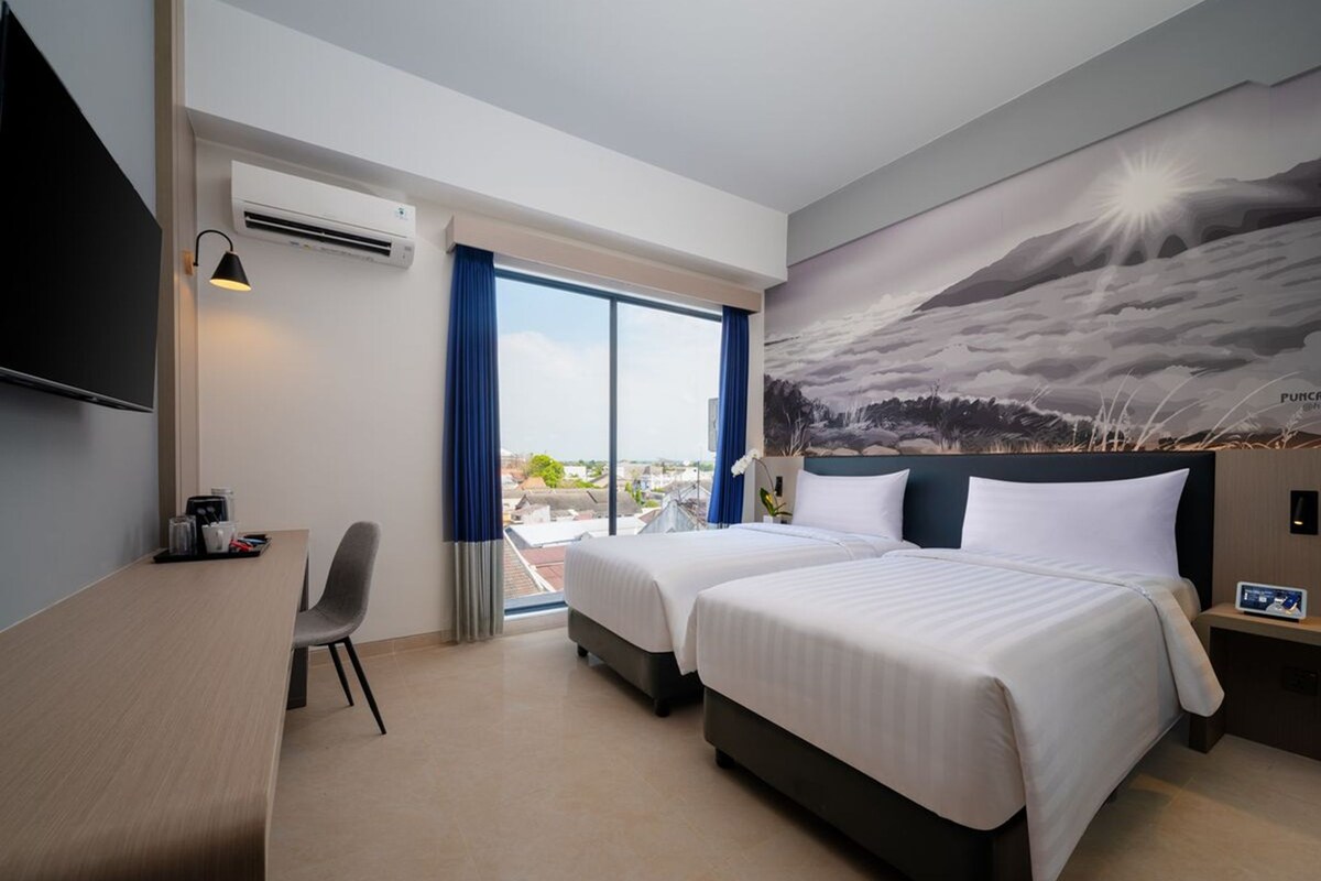 A hotel room with two beds, a desk, a chair, a TV, and a large window with a view of the city.