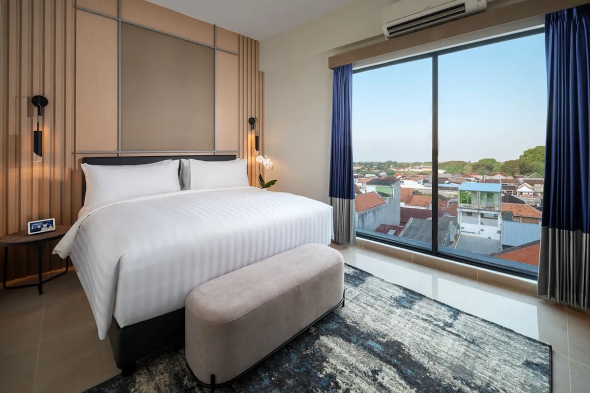 A modern hotel room with a large bed, a bench, a nightstand, an air conditioner, and a large window with a city view.