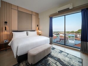 A modern hotel room with a large bed, a bench, a nightstand, an air conditioner, and a large window with a city view. - Aston Inn Lumajang - East Java (Lumajang)