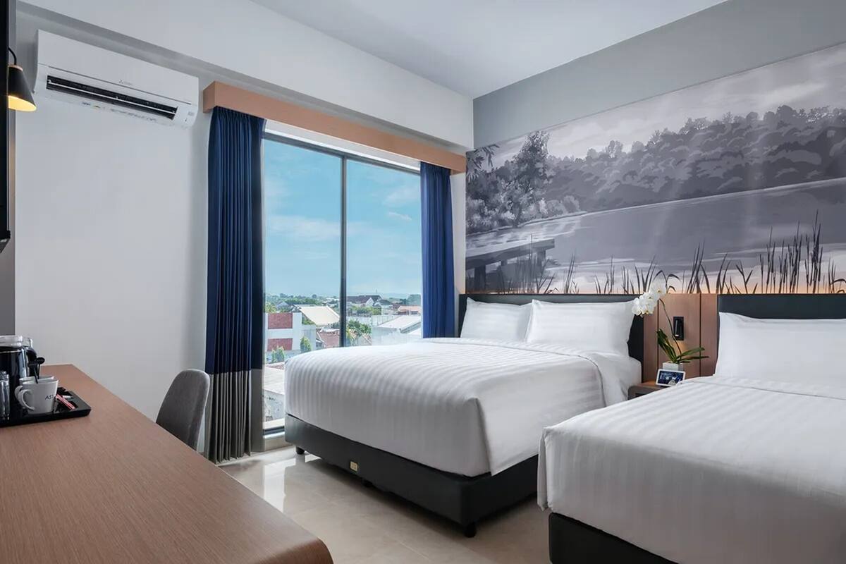 Habitación de hotel con dos camas, un escritorio y un ventanal grande que ofrece vistas a la ciudad.