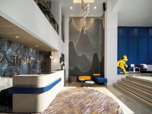 A modern hotel lobby with a reception desk, a seating area with a sofa and ottoman, a staircase, and a decorative wall. - Aston Inn Lumajang - East Java (Lumajang)