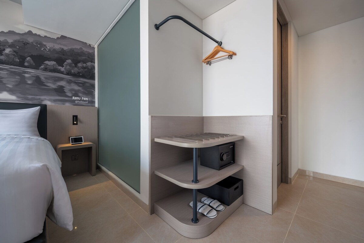 A modern hotel room with a curved corner design, a bed, a small bedside table with a lamp, a wall-mounted radio, and a shoe storage unit.