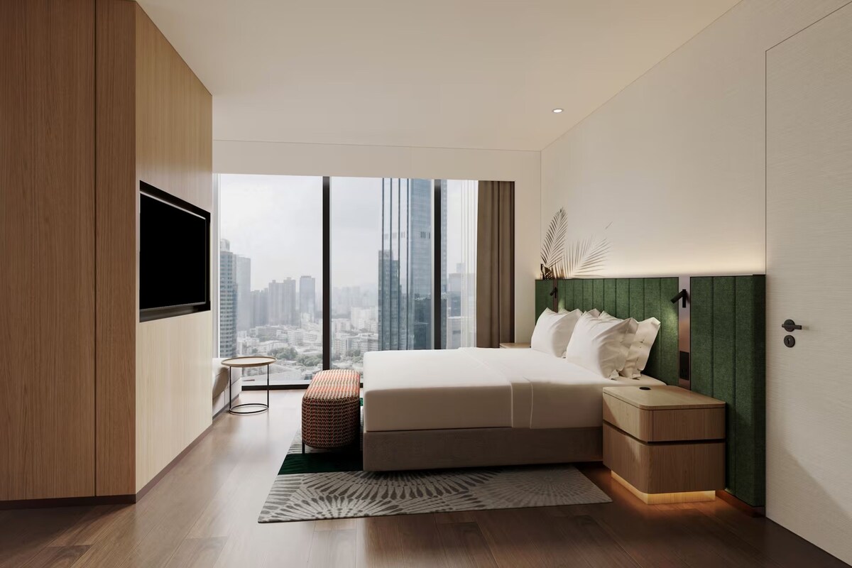 A modern bedroom with a large bed, a TV mounted on the wall, a nightstand, and a view of the cityscape.
