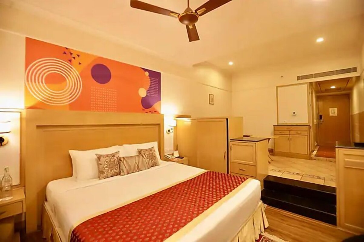 A hotel room with a bed, a ceiling fan, a nightstand, a mirror, and a small kitchenette.