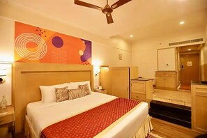 A hotel room with a bed, a ceiling fan, a nightstand, a mirror, and a small kitchenette. - Ira By Orchid Mumbai - T2 International Airport (Mumbai)