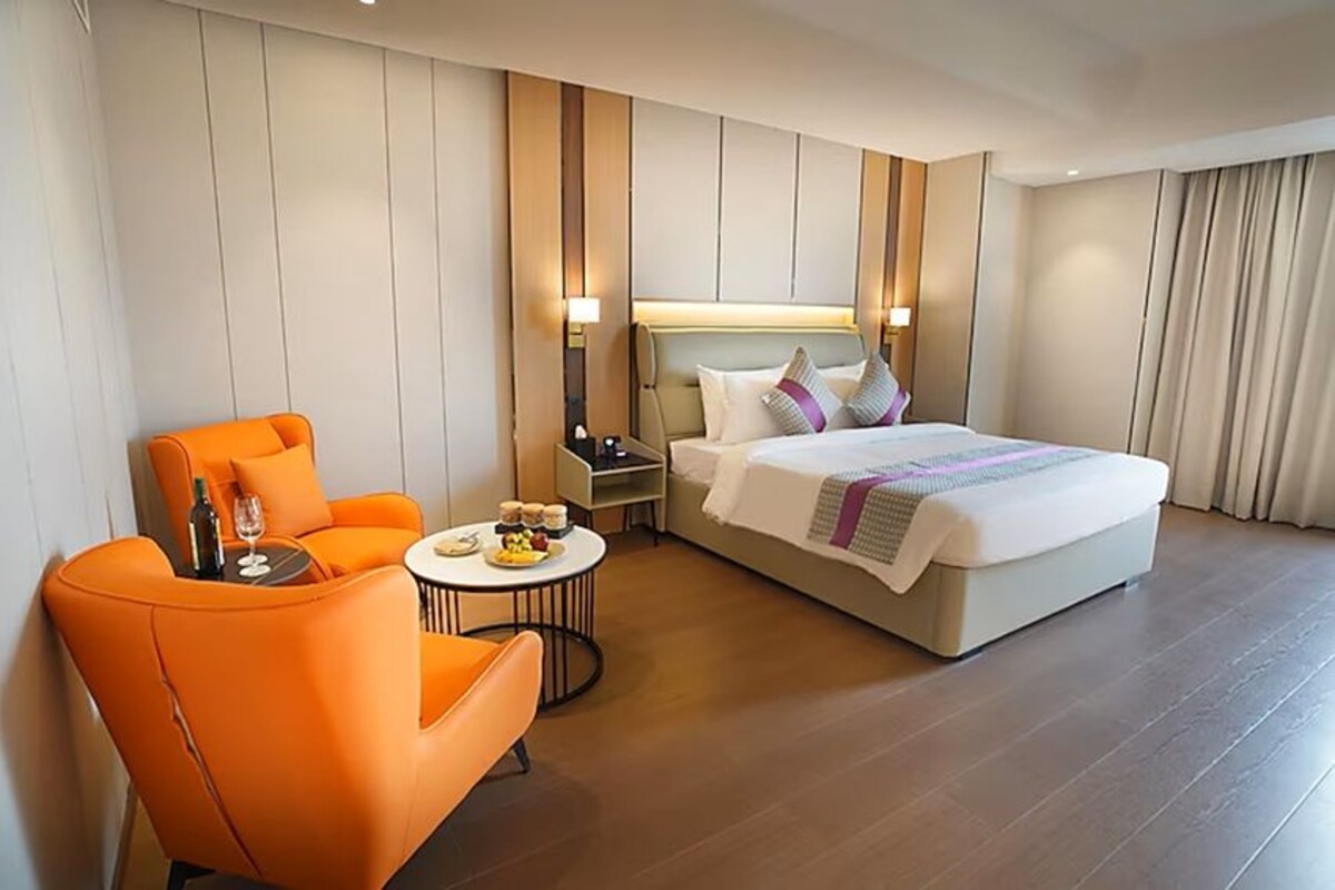 A hotel room with a bed, an orange armchair, a small round table with a bottle and glasses, and a nightstand with a lamp.