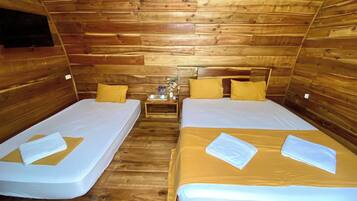 A cabin with two wooden beds, each with a mattress and pillows. There is a small wooden nightstand with a cup, a bottle, and a small table with a vase.