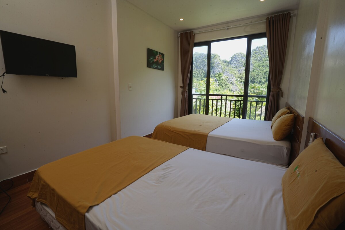 Ein Hotelzimmer mit zwei Betten, einem an der Wand montierten Fernseher, einem Fenster mit Vorhängen und einem Balkon mit Blick auf Grünflächen.