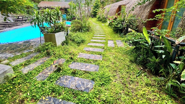 A pathway leading to a pool surrounded by lush greenery and thatched-roof structures. - Catba Backpackers Hostel & Pool Bar (Hai Phong)