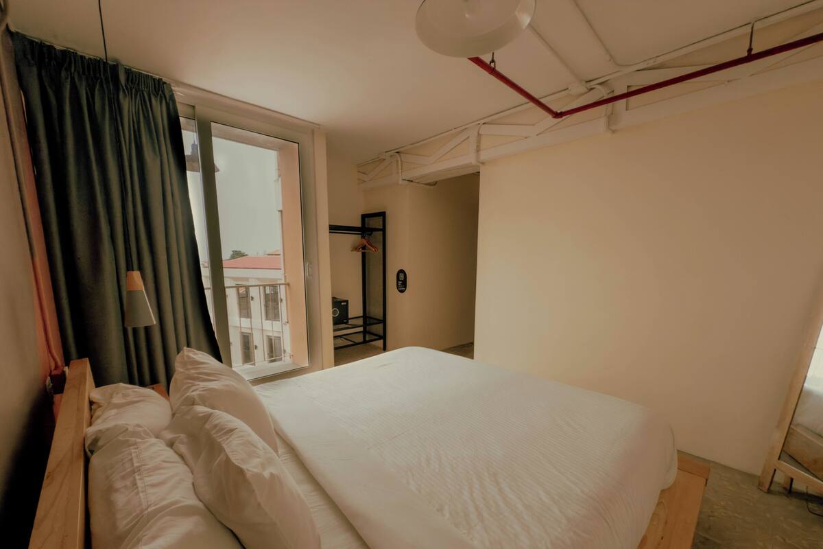 Een slaapkamer met een groot bed, een raam met gordijnen, een hanglamp en uitzicht op een gebouw.