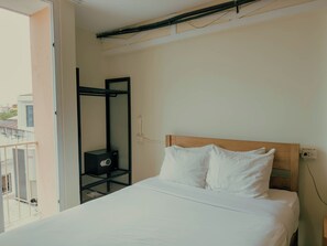 Una cama individual con ropa blanca, cabecera de madera y una mesita de noche con enchufe. Un altavoz negro está montado en un estante de metal negro adosado a la pared.