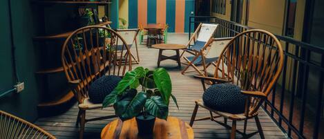 Una terraza de madera con sillas de mimbre y una mesa central, rodeada de plantas en macetas.
