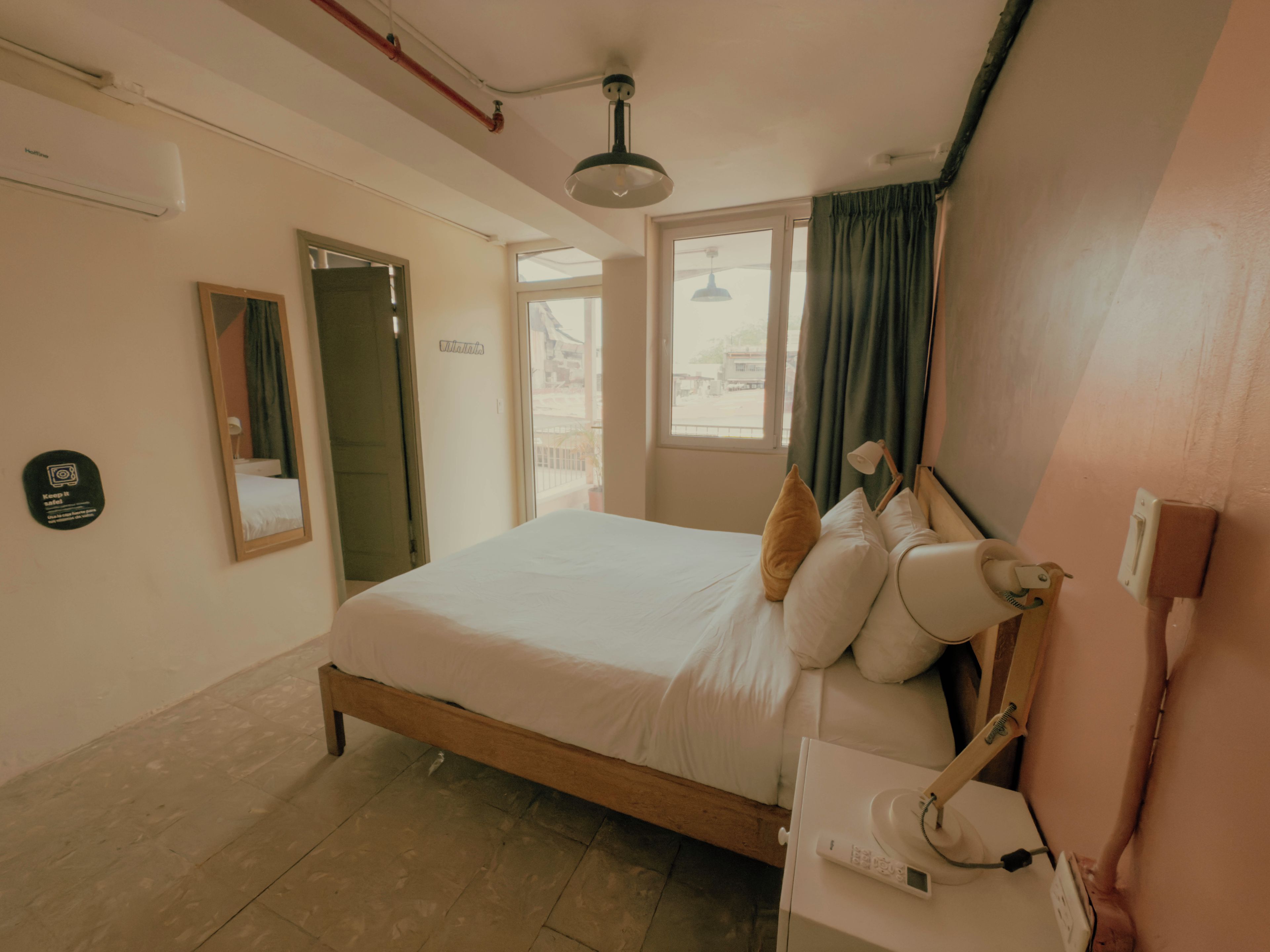 A hotel room with a bed, bedside table, lamp, and a view of the outside through a window.