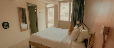 A hotel room with a bed, bedside table, lamp, and a view of the outside through a window.