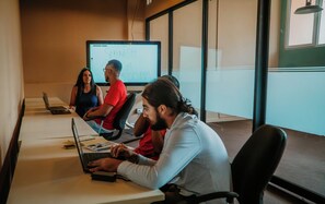 Un salón de reuniones con una pantalla grande mostrando una presentación, una computadora portátil y participantes sentados alrededor de una mesa.