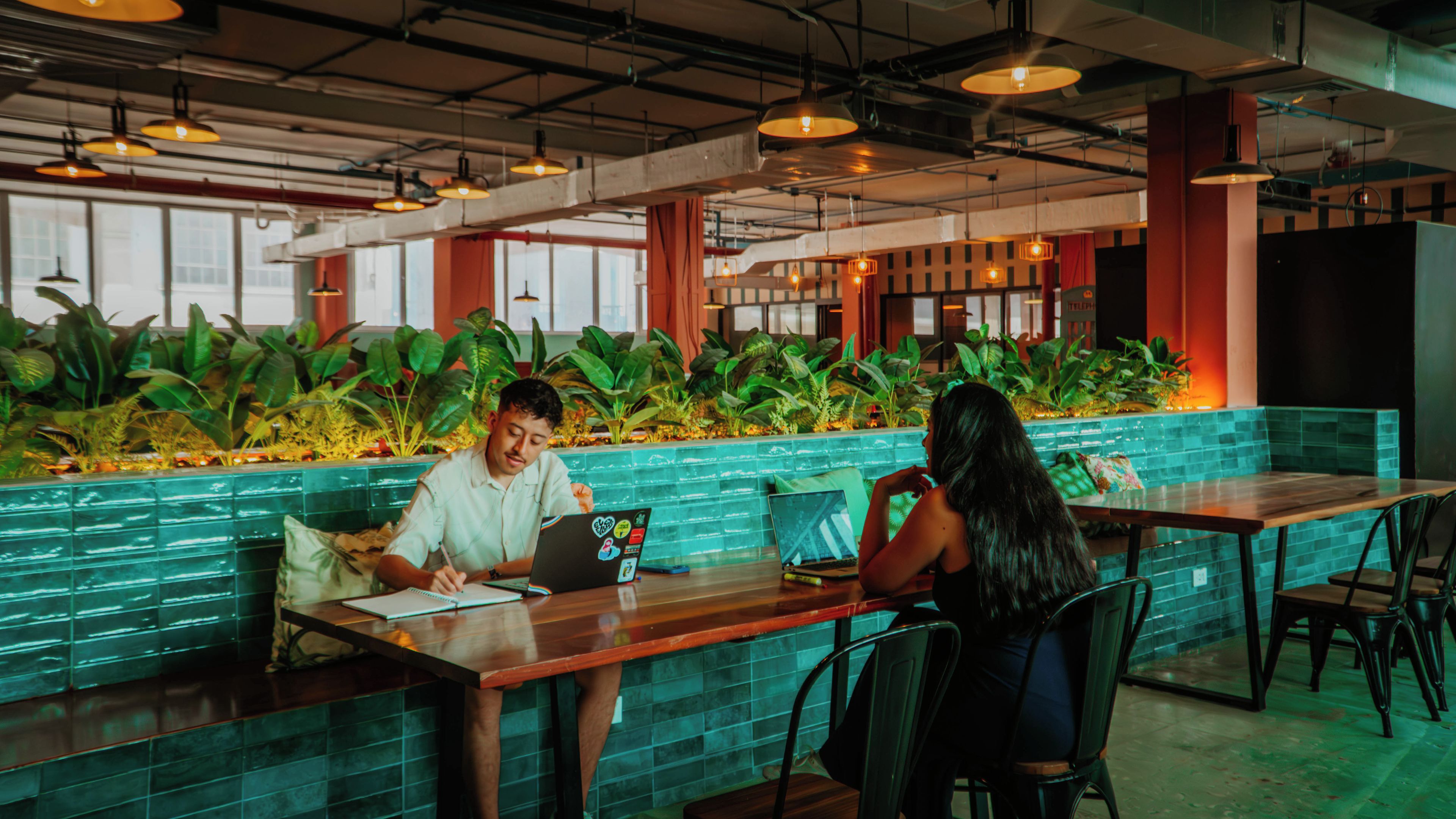A modern indoor space with a long counter, two people seated at a table, and a backdrop of green plants.