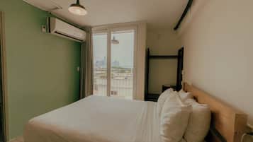 Una habitación de hotel con cama, ventana con vistas a un paisaje urbano y un equipo de aire acondicionado en la pared.