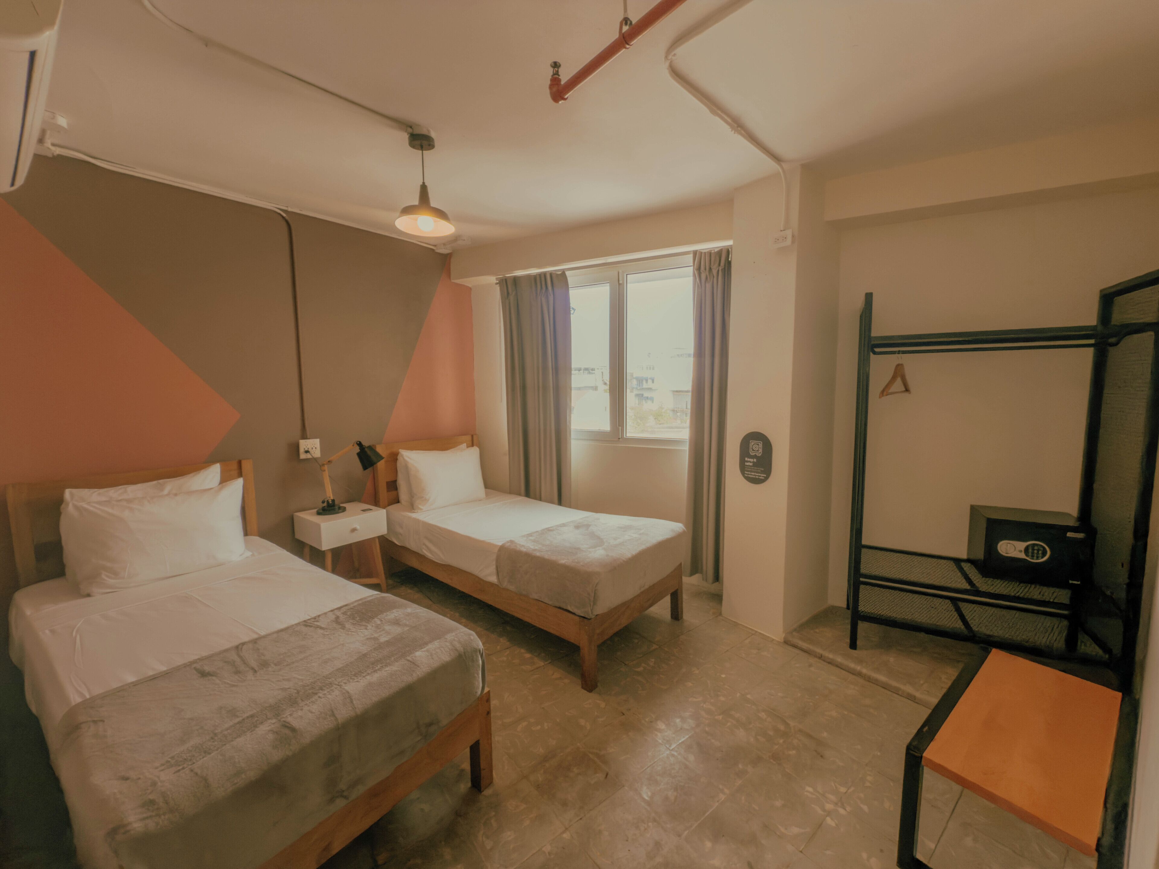 A hotel room with two single beds, a desk with a chair, a window with curtains, and a view of the outside.