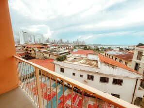Un edificio alto con techo de tejas rojas, un balcón con barandilla de metal y vista a un paisaje urbano con un puente a lo lejos.